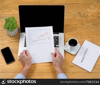 High angled view of male hands holding data graph with computer, coffee, notepad, pen and plant on desktop.&#xA;&#xA;