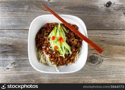 High angled view of noodles with ground beef and cucumbers. Chopsticks on top of bowl with rustic wood underneath.