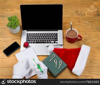 High angled view of Santa Claus writing his gift list with hot chocolate, computer, present, cap, notepad, pencil, cell phone, reading glasses and plant on desktop. Christmas concept of Santa Claus office.