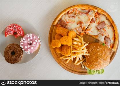 High calorie food on the table, top view, nobody. Pizza and burger, doughnuts, french fries and chicken nuggets. Junk fastfood