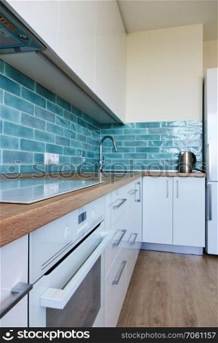 High glossy, white kitchen with blue tile, brown top, modern hob and oven. White glossy kitchen with modern appliances