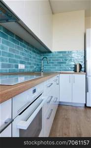 High glossy, white kitchen with blue tile, brown top, modern hob and oven. White glossy kitchen with modern appliances