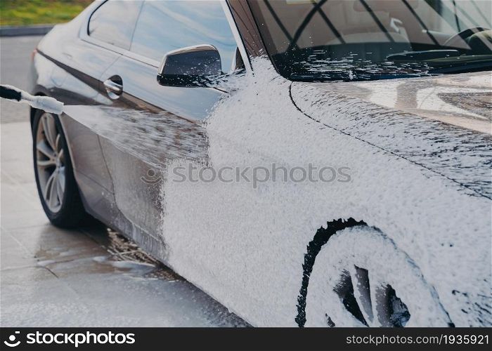 High-pressure car cleaning, worker at car wash station spraying special wash soap on vehicle with pressure washer outdoors, auto surface in white foam during manual exterior wash. High-pressure car cleaning, spraying special wash soap on vehicle with pressure washer outdoors