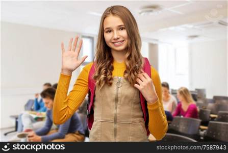high school, education and people concept - smiling teenage student girl with backpack waving hand over lecture hall background. happy teenage student girl with backpack at school