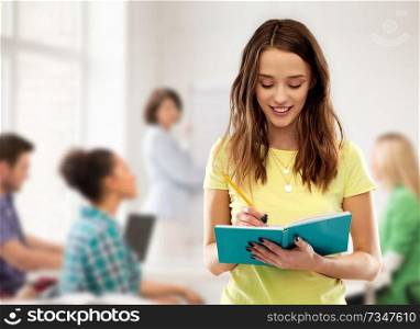 high school, education and people concept - smiling young teenage student girl in yellow t-shirt writing to diary or notebook by pencil over classroom and teacher background. teenage student girl writing to notebook at school