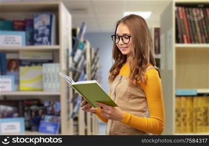 high school, education and vision concept - happy smiling teenage student girl in glasses reading book over library background. student girl in glasses reading book at library
