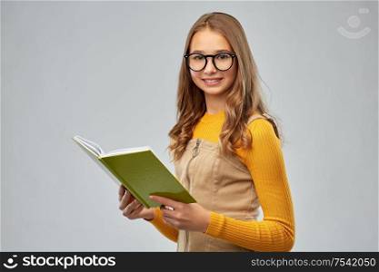 high school, education and vision concept - smiling teenage student girl in glasses reading book over grey background. teenage student girl in glasses reading book