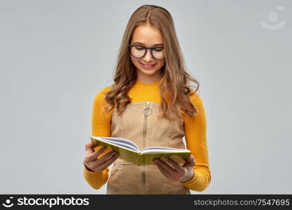 high school, education and vision concept - smiling teenage student girl in glasses reading book over grey background. teenage student girl in glasses reading book