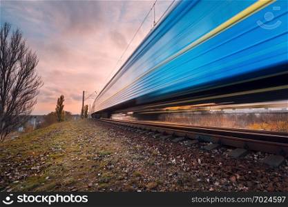 High speed blue passenger train on railroad track in motion at sunset. Blurred commuter train. Railway station with cloudy sky. Railroad travel, railway tourism. Industrial landscape at dusk