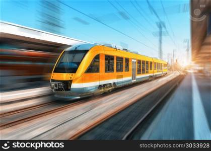High speed orange train in motion on the railway station at bright day. Modern intercity train with motion blur effect on the railway platform. Industrial. Passenger commuter train on railroad. Travel. High speed orange train in motion on the railway station