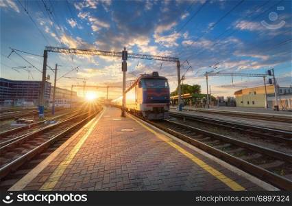 High speed passenger train in motion on railroad track. High speed passenger train on railroad track at sunset in Europe. Railway station with modern commuter train departure, blue sky with clouds and sun. Colorful Industrial landscape. Railway platform