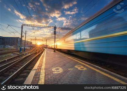 High speed passenger train in motion on railroad track. High speed passenger train on railroad track at sunset in Europe. Railway station with modern commuter train departure, blue sky with clouds and sun. Colorful Industrial landscape. Railway platform