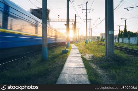 High speed passenger train in motion on railroad track at sunset. Railway station with blurred modern commuter train in the evening in Europe. Industrial landscape with path, train and green grass