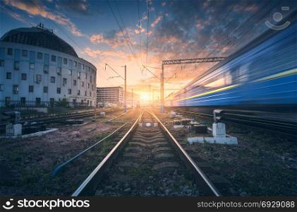 High speed passenger train in motion on railroad track at sunset. Railway station with blurred modern commuter train, against colorful blue sky with red and orange clouds at dusk.Industrial landscape