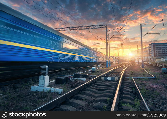 High speed passenger train in motion on railroad track at sunset. Railway station with blurred modern commuter train, against colorful blue sky with red and orange clouds at dusk.Industrial landscape