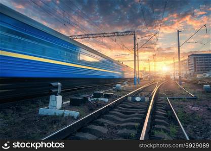 High speed passenger train in motion on railroad track at sunset. Railway station with blurred modern commuter train, against colorful blue sky with red and orange clouds at dusk.Industrial landscape