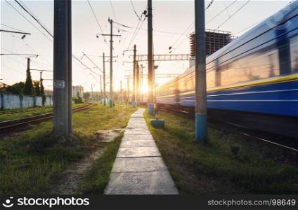 High speed passenger train in motion on railroad track at sunset. Railway station with blurred modern commuter train, railway traffic light against colorful blue sky with clouds. Industrial landscape
