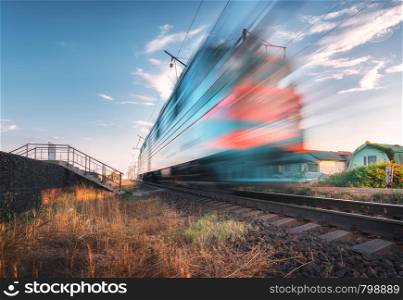 High speed passenger train in motion on the railroad at summer evening. Moving blurred modern commuter train at sunset. Industrial landscape with railway station and blue sky. Transport. Travel. High speed passenger train in motion on the railroad at sunset
