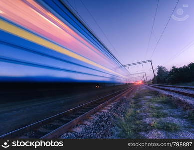 High speed passenger train in motion on the railroad at summer night. Moving blurred modern commuter train at dusk. Industrial landscape with railway station and purple sky. Transport. Travel. High speed passenger train in motion on the railroad at night