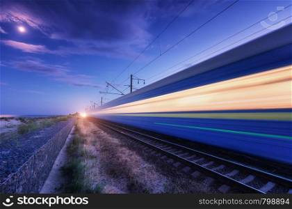 High speed passenger train in motion on the railroad at summer night. Moving blurred modern commuter train at dusk. Railway station and purple sky with moon and clouds. Industrial landscape. Transport. High speed passenger train in motion on the railroad at night