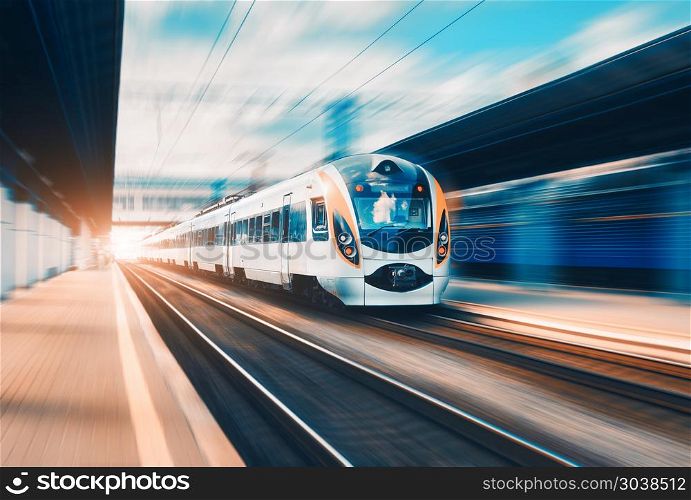 High speed passenger train in motion on the railway station at sunset in Europe. Modern intercity train on railway platform with motion blur effect. Urban scene with railroad. Railway transportation. High speed train at the railway station at sunset