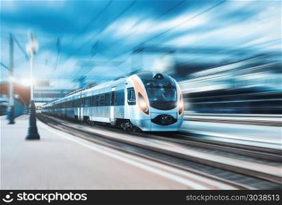 High speed passenger train in motion on the railway station at sunset in Europe. Modern intercity train on railway platform with motion blur effect. Urban scene with railroad. Railway transportation. High speed train at the railway station at sunset
