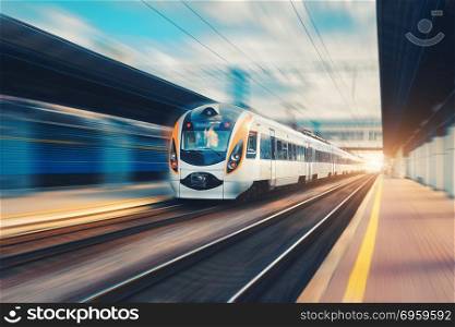 High speed passenger train in motion on the railway station at sunset in Europe. Modern intercity train on railway platform with motion blur effect. Urban scene with railroad. Railway transportation. High speed passenger train in motion on railroad