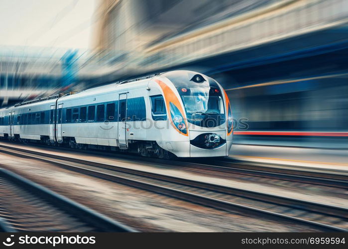 High speed passenger train in motion on the railway station at sunset in Europe. Modern intercity train on railway platform with motion blur effect. Urban scene with railroad. Railway transportation. High speed passenger train in motion on railroad