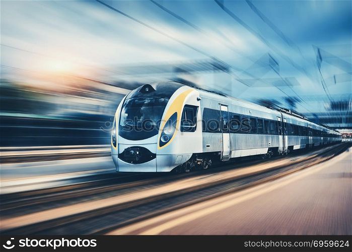 High speed passenger train in motion on the railway station at sunset in Europe. Modern intercity train on railway platform with motion blur effect. Urban scene with railroad. Railway transportation. High speed passenger train in motion on railroad