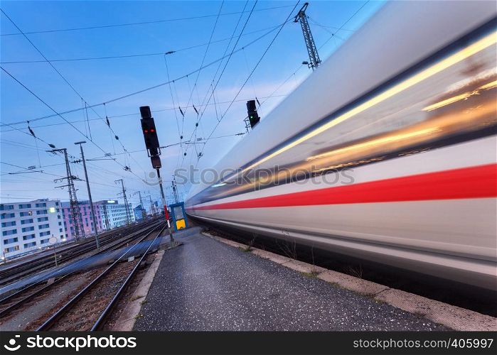 High speed passenger train on railroad track in motion at night. Blurred commuter train. Railway station at twilight in Nuremberg, Germany. Railroad travel, railway tourism. Industrial landscape