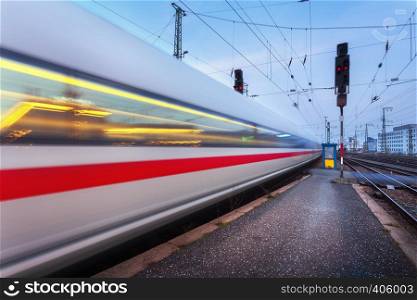 High speed passenger train on railroad track in motion at night. Blurred commuter train. Railway station at twilight in Nuremberg, Germany. Railroad travel, railway tourism. Industrial landscape