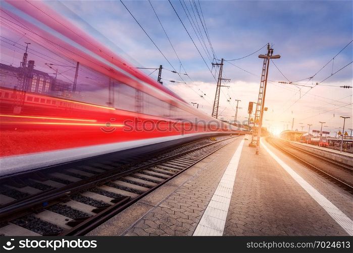High speed red passenger train on railroad track in motion at beautiful sunset. Blurred commuter train. Railway station in the evening. Railroad travel, railway tourism. Industrial landscape. Train