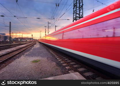 High speed red passenger train on railroad track in motion at sunset. Blurred commuter train. Railway station in Nuremberg, Germany. Industrial landscape