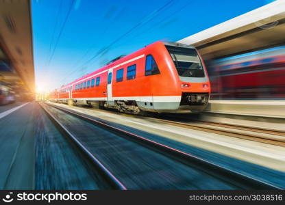 High speed red train in motion on the railway station at sunset. Modern intercity vehicle with motion blur effect on the railway platform. Industrial. Passenger commuter train on railroad. Travel. High speed red train in motion on the railway station