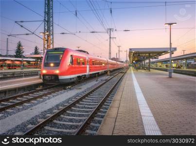 High speed red train on the railway station at sunset. Nuremberg, Germany. Colorful urban landscape. Modern intercity train on the railway platform at night. Commuter passenger train on railroad. Dusk. High speed red train on the railway station at dusk
