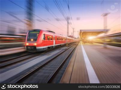 High speed red train with motion blur effect on the railway station at sunset. Landscape. Modern intercity train in motion on the railway platform at night. Commuter passenger train on railroad. Dusk. High speed red train with motion blur effect on the railway station