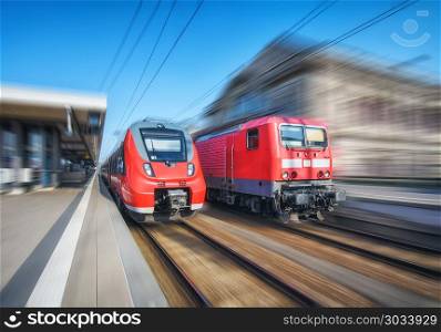 High speed train and old train in motion on the railway station at sunset. Modern intercity train on the railway platform. Industrial landscape. Commuter red trains with motion blur effect on railroad. High speed train and old train in motion on the railway station