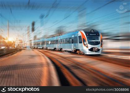 High speed train arrives on the railway station at sunset. Modern intercity train in motion on the railway platform. Passenger train on railroad with motion blur effect. Railway transportation