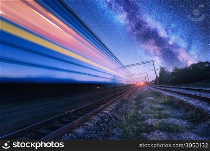 High speed train in motion and Milky Way at starry night. Industrial landscape with sky and stars over blurred modern passenger train and railroad. Railway station and space. Technology and nature