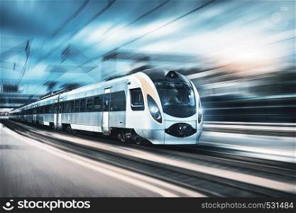 High speed train in motion at the railway station at sunset in Europe. Modern intercity train on the railway platform with motion blur effect. Industrial landscape with passenger train on railroad