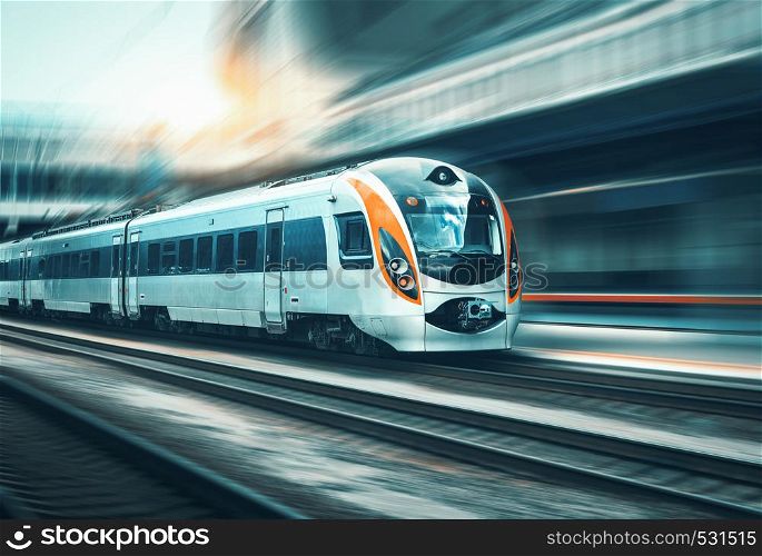 High speed train in motion at the railway station at sunset in Europe. Modern intercity train on the railway platform with motion blur effect. Industrial landscape with passenger train on railroad. High speed train in motion at the railway station