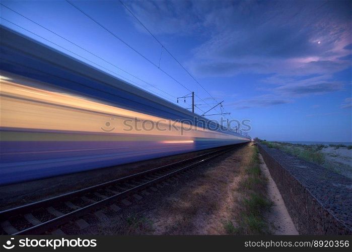 High speed train in motion on the railway station at dusk. Landscape with moving blue modern intercity passenger train, rural railway platform, blue sky. Railroad in europe. Railway transportation