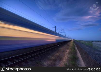 High speed train in motion on the railway station at dusk. Landscape with moving blue modern intercity passenger train, rural railway platform, blue sky. Railroad in europe. Railway transportation