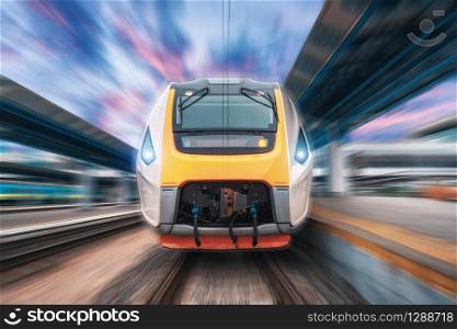 High speed train in motion on the railway station at sunset. Fast moving modern passenger train on the railway platform. Railroad with motion blur effect. Commercial transportation. Front view
