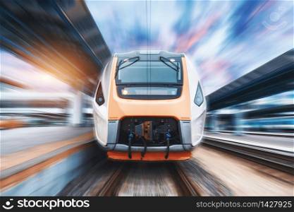 High speed train in motion on the railway station at sunset. Fast moving modern passenger train on the railway platform. Railroad with motion blur effect. Commercial transportation. Front view. High speed train in motion on the railway station. Front view.