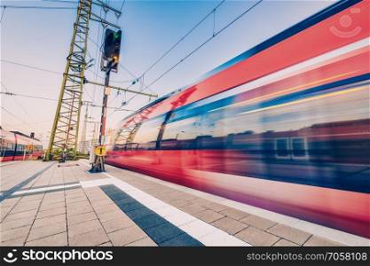 High speed train in motion on the railway station at sunset. Red blurred modern intercity train on the railway platform in Europe. Passenger railway transportation. Railroad in the evening. Travel