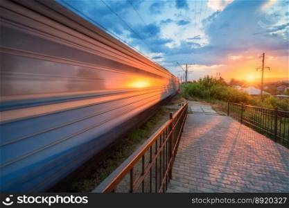 High speed train in motion on the railway station at sunset in summer. Moving modern intercity passenger train, railway platform, green trees and sunlight. Railroad in Urkaine. Railway transportation. High speed train in motion on the railway station at sunset