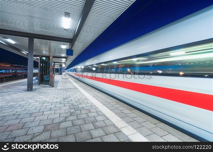 High speed train in motion on the railway station with beautiful illumination at night. Moving blurred modern intercity train on the railway platform. Passenger transportation. Railroad. Travel. High speed train in motion on the railway station at night