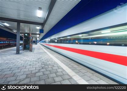 High speed train in motion on the railway station with beautiful illumination at night. Moving blurred modern intercity train on the railway platform. Passenger transportation. Railroad. Travel. High speed train in motion on the railway station at night