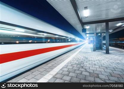 High speed train in motion on the railway station with beautiful illumination at night. Moving blurred modern intercity train on the railway platform. Passenger transportation. Railroad. Travel